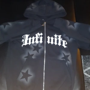 Svart hoodie med stjärnor - Svart hoodie med vit text och stora stjärnor i sprayad stil över hela plagget. Dragkedja framtill med stjärnformad dragkedjetag. Huva och känguruficka. Avslappnad passform, perfekt för streetwear. 10/10 skick helt ny. XS men passar även S