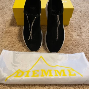 Svarta sneakers från Diemme med dragkedja - Säljer ett par svarta sneakers från Diemme med vit sula och dragkedja framtill. Skorna har en modern design med rund tå och är tillverkade i mocka. Perfekta för dig som vill ha något stilrent men ändå unikt. Kvitto kan visas på begäran.