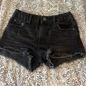 Svarta jeansshorts med fransar från Gina Tricot - Snygga svarta jeansshorts från Gina Tricot i samarbete med @izaandelle. Shortsen har fransade benslut, klassisk femficksdesign och knappgylf. Perfekta för sommaren och ger en cool, avslappnad look.