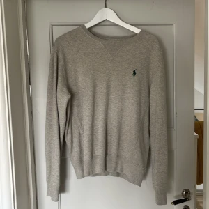 Grå sweatshirt från Polo Ralph Lauren - Klassisk grå sweatshirt från Polo Ralph Lauren med rund halsringning och broderad grön logga på bröstet. Tröjan har långa ärmar och ribbade muddar vid ärmslut och nederkant. 