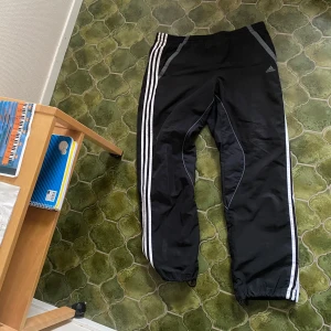 Svarta träningsbyxor från Adidas - Svarta träningsbyxor från Adidas med vita ränder längs sidorna och grå detaljer vid fickorna. Byxorna har resår i midjan och vid bensluten, samt Adidas-logga på vänster sida.