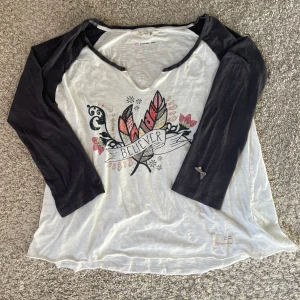 Vit och svart långärmad topp med tryck - Säljer en vit topp med svarta ärmar och v-ringning från Odd Molly. Framsidan har ett färgglatt fjäder- och texttryck med ordet 'Believer'. Ärmarna är trekvarts och materialet känns mjukt och tunt. Detaljerna som kan uppfattas som "klippta" vid slutet av tröjan och i halsringningen är en del av designen. 