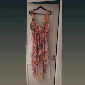 Blommig omlottklänning - Säljer en vacker blommig omlottklänning i ljusa toner av orange, blått och beige. Klänningen har en v-ringad design och är ärmlös, perfekt för sommarens festligheter. Den har ett fint fall och ett elastiskt band i midjan för en bekväm passform.