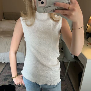 Vit ärmlös topp med struktur - Säljer en vit ärmlös topp med hög hals och strukturerat tyg. Toppen har en rak passform och är tillverkad i ett lätt, syntetiskt material. Perfekt för finare tillfällen! Använd en gång🤍🤍