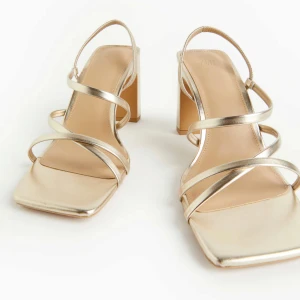 Guldiga sandaler med klack och smala remmar - Säljer ett par trendiga sandaler i guld med fyrkantig tå och breda klackar. Skorna har smala remmar som korsar foten. Köpta för balen men var för små för mig. Aldrig använda. 