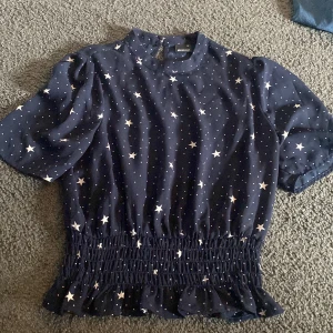 Mörkblå blus med stjärnmönster från Gina Tricot - Supersöt mörkblå blus från Gina Tricot med vita stjärnor och prickar. Blusen har korta puffärmar och smockad midja med volangkant nedtill. Storlek 36 och säljs då den inte används. 