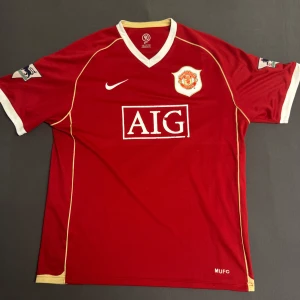 Manchester United fotbollströja Ronaldo 7 - Klassisk röd Manchester United fotbollströja med vita och gula detaljer, AIG sponsortryck och klubbmärke på bröstet. Nummer 7 och Ronaldo på ryggen. Kortärmad modell med V-ringning och broderade loggor på ärmarna. Lite slitage på sponsorn. 