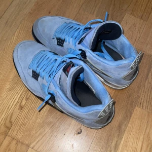 Nike Air Jordans 4 University Blue - Säljer ett par Nike Air Jordans 4 University Blue med ljusblå mocka och matchande blå skosnören. Skorna har svarta och grå detaljer samt den klassiska Jumpman-loggan på hälen. Perfekta för dig som vill sticka ut med snygga sneakers.