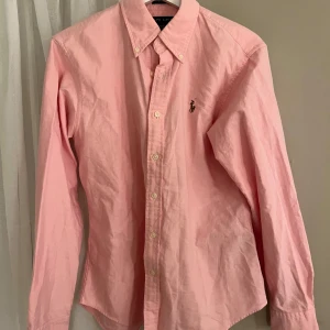 Rosa skjorta från Ralph Lauren - Klassisk rosa skjorta från Ralph Lauren med button-down krage och broderad logga på bröstet. Skjortan har långa ärmar och vita knappar framtill. Perfekt för en stilren och avslappnad look.