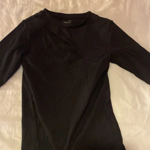 Svart ribbad topp med omlottdetalj - Säljer en svart ribbad topp med omlottdetalj vid halsen. Toppen har trekvartsärm och är i ett stretchigt material som sitter snyggt på kroppen. Perfekt att styla till både jeans och kjol.