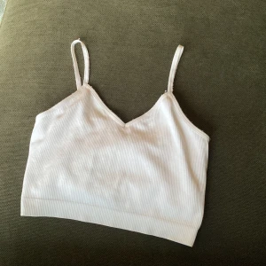 Vit ribbad linnetopp från Primark - Säljer en vit ribbad linnetopp från Primark med smala axelband och v-ringning. Toppen är i mjuk och stretchig polyesterblandning, perfekt för varma dagar eller som lager under andra plagg.
