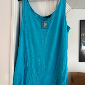 Turkos linne med pärldetaljer från Lindex - Säljer en turkos ärmlös linne från Lindex i storlek L. Klänningen har dekorativa pärlor längs halsringningen och ett fyrkantigt pärlbroderi framtill. Saknas pärlor/stenar på vissa ställen och ett litet hål bak på baksidan (se bild)
