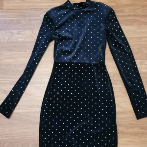 Svart långärmad klänning med pärlor - Snygg svart långärmad klänning med små pärlor över hela tyget. Hög hals och figurnära passform, perfekt för dig som vill sticka ut lite extra. Klänningen har en elegant och stilren look.