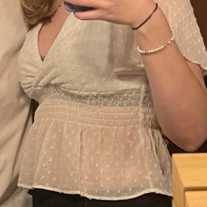 Vit blus med omlott och volang - Säljer en söt vit blus med omlott framtill och volang nertill. Blusen har korta ärmar och är tillverkad i ett lätt, transparent material med små prickar i tyget. Smockad midja för en fin passform.