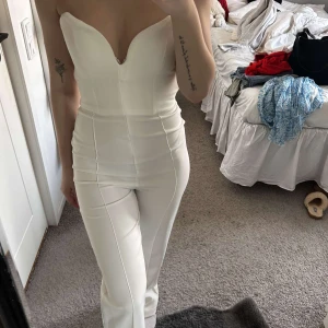 Vit byxdress med sweetheart-urringning -  elegant vit byxdress med figurnära passform och axelbandslös sweetheart-urringning. Byxdressen har raka ben och markerad midja, perfekt för dig som vill sticka ut med en stilren look. Den är superfin till finare tillfällen och sitter superfint på kroppen! Inte använd 