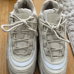 Beige sneakers från Fila - Säljer ett par snygga beige sneakers från Fila med vita detaljer. Skorna har snörning och en chunky sula som ger en sportig look. Perfekta för dig som gillar streetwear och vill ha bekväma skor till vardags.