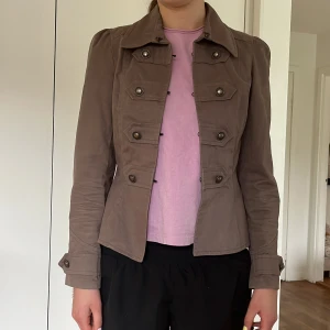 Brun kavajjacka från Gosha - Snygg brun kavajjacka från Gosha med markerad midja och dekorativa knappar framtill. Jackan har klassisk krage, lång ärm och flera fickor med lock. Perfekt för dig som vill ha en stilren och tidlös look.