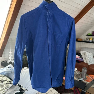 Blå skjorta från These Glory Days - Säljer en stilren blå skjorta från These Glory Days med ståkrage och bröstficka. Skjortan har långa ärmar och knäppning framtill. Perfekt för dig som gillar en enkel men snygg look. Storlek M