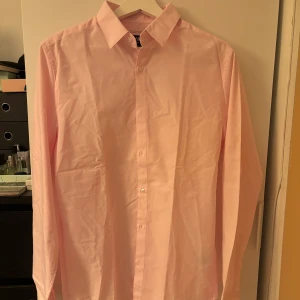 Ljusrosa skjorta från H&M - Säljer en ljusrosa skjorta från H&M i slim fit-modell. Skjortan har klassisk krage, långa ärmar och knäppning framtill. Tillverkad i en blandning av polyester och bomull som gör den lätt att stryka. Perfekt för dig som gillar stilrena plagg.