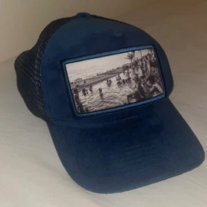 Mörk blå keps från Emilio Pendenza -  mörkblå keps med blått nät baktill och ett fototryck framtill som föreställer en badplats. Kepsen har böjd skärm och justerbar passform. Perfekt accessoar för en avslappnad stil. 