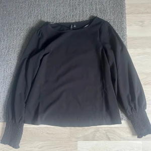 Svart blus från Vero Moda - Säljer en svart blus från Vero Moda med långa ärmar och smockade muddar vid ärmsluten. Blusen har en rak passform. Säljer för jag ej använder.