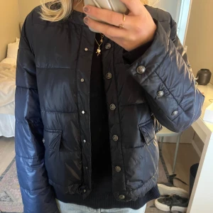 Mörkblå pufferjacka med knappar från Massimo Dutti - Säljer min mörkblå pufferjacka med rund halsringning och dekorativa silverfärgade knappar framtill. Jackan har två fickor med lock och är långärmad. Perfekt för kyligare dagar och enkel att matcha med olika outfits. 