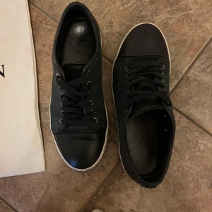 Svarta sneakers från Lanvin med krokodilmönster - Säljer ett par svarta sneakers från Lanvin med snygg krokodilpräglad yta och vita sulor. Skorna har rund tå, snörning och är tillverkade i skinn. Perfekta för dig som vill ha en stilren och lyxig look.
