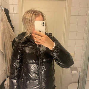 Svart pufferjacka Nelly - Säljer en svart, glansig pufferjacka med huva och dragkedja framtill. Jackan har en klassisk pufferdesign och är perfekt för kyligare dagar. Snygg och enkel att matcha till olika outfits.