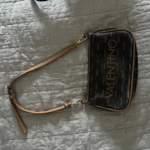 Svart axelväska från Valentino - Säljer en brun axelväska från Valentino med guldiga logotexter och detaljer. Väskan har justerbart beige axelband och dragkedja upptill. Perfekt för att bära det viktigaste och ge en lyxig touch till din outfit.