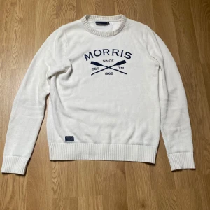 Vit stickad tröja från Morris - Vit stickad tröja från Morris med rund halsringning och blå logotyptryck framtill. Tröjan har ribbade muddar vid ärmslut och nederkant, vilket ger en klassisk och stilren look.