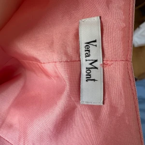 Aftonklänning  från Vera Mont - Säljer en elegant rosa långklänning från Vera Mont med v-ringning och smala axelband. Klänningen har en figurnära överdel och utställd kjol i ett glansigt material som ger en lyxig känsla. Perfekt för speciella tillfällen.