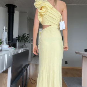 Ljusgul långklänning med volang och oneshoulder - Säljer en ljusgul långklänning med oneshoulder och stor volangdetalj över axeln. Klänningen har en elegant skärning med utskuren midja och figurnära passform. Perfekt för bal eller festliga tillfällen.
