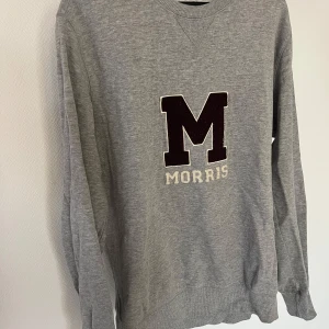 Grå sweatshirt från Morris - Säljer en klassisk grå sweatshirt från Morris med stor mörklila M och broderad text på bröstet. Tröjan har rund halsringning och långa ärmar. Perfekt för en avslappnad och stilren look.