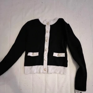 Svart och vit kofta från Amisu - Säljer en svart kofta från Amisu med vita detaljer vid krage, knappslå, nederkant och fickor. Koftan har guldknappar framtill och är långärmad. Perfekt för dig som gillar klassisk och stilren design. Vi kan diskutera priset😊