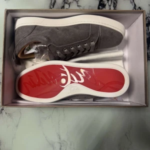 Louboutins - Säljer ett par gråa sneakers från Christian Louboutin i mocka med vit sula och den klassiska röda undersidan. Skorna har snörning och rund tå, perfekt för dig som vill sticka ut med exklusiva detaljer.