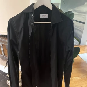 Svart skjorta från Eton - Säljer en svart skjorta från Eton i modellen super slim. Skjortan har klassisk krage och knäppning framtill. Perfekt för dig som gillar en stilren och tajt passform.