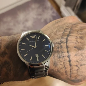 Emporio Armani herrklocka med mörkblå urtavla - Stilren klocka från Emporio Armani med mörkblå urtavla, silverfärgade visare och index samt datumvisning. Boett och länk i silverfärgat metall. Perfekt accessoar för dig som gillar klassisk design.