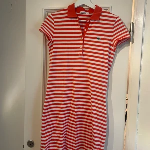 Röd- och vitrandig pikéklänning från Lacoste - Säljer en klassisk pikéklänning från Lacoste i röd- och vitrandigt mönster. Klänningen har krage, knappar framtill och korta ärmar. Perfekt för en sportig och avslappnad look. Ikoniska Lacoste-loggan på bröstet.