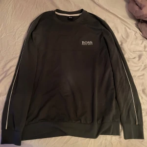 Svart sweatshirt från Hugo Boss - Säljer en svart sweatshirt från Hugo Boss med vit logga broderad på bröstet. Tröjan har långa ärmar med vita ränder längs sidorna och ribbade muddar vid ärmslut och nederkant. Perfekt för en stilren och avslappnad look.