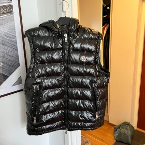 Svart dunväst från Moncler - Snygg svart dunväst från Moncler med glansig finish och huva. Västen har dragkedja framtill, två fickor med dragkedja och Moncler-logga på bröstet. Perfekt för lager-på-lager och stilren look. Storlek L.