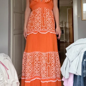 Maxiklänning  - Orange långklänning med vita broderade blommor och volangdetaljer. Djup v-ringning och volangkant upptill. Använd en gång och i jättebra skick. Som man kan se på sista bilden finns det lite svarta fläckar. Dem är på insidan av ryggen och syns inte och går säkert bort i tvätt. Jag är 175 cm lång som referens 
