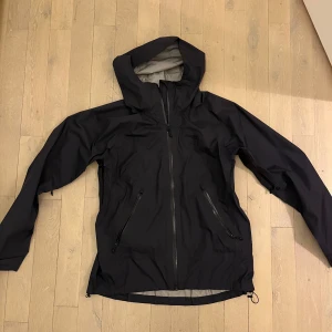  vind/regnjacka med huva Bergans - Säljer en svart vindjacka med huva och dragkedja framtill. Jackan har två snedställda fickor med dragkedja och justerbar nederkant. Perfekt för blåsiga dagar och enkel att matcha med det mesta.