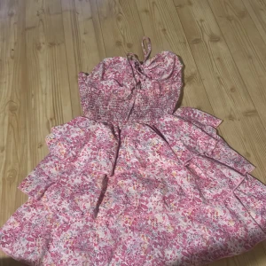 Rosa blommig axelbandslös klänning med volanger - Söt axelbandslös klänning i rosa och vita toner med blommigt mönster och volangdetaljer. Smockad överdel och knytband upptill. Perfekt för sommarens varma dagar.