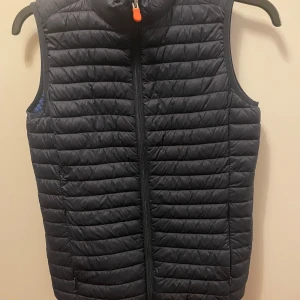 Navy blue  dunväst från Save The Duck - Säljer en navy blue dunväst från Save The Duck med horisontella sömmar och dragkedja framtill. Västen har en hög krage och en orange logotypdetalj. Perfekt för lager-på-lager och tillverkad i syntetmaterial.