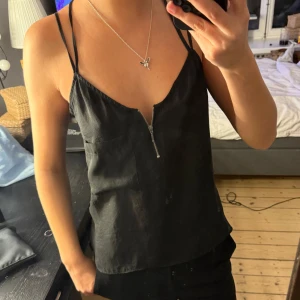 Svart linnetopp med dragkedja - Snygg svart linnetopp med smala axelband som korsas i ryggen och en liten dragkedjedetalj framtill.