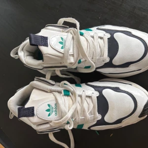 Vita och marinblå sneakers från Adidas - Säljer ett par snygga Adidas sneakers i vitt och marinblått med gröna detaljer. Skorna har klassisk Adidas-logga på plösen och vita snören. Perfekta för dig som gillar retrostil och streetwear.