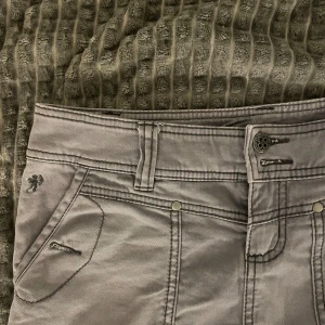 Grå jeanskjol med rå kant - Säljer en grå kort jeanskjol med rå, fransig nederkant och dekorativa sömmar. Kjolen har två framfickor med dragkedja och två bakfickor med knappdetaljer. Perfekt för dig som gillar en cool och avslappnad stil.