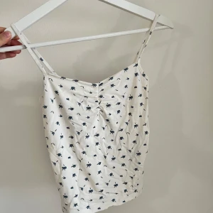 Vit blommig linne med smala axelband - Söt vit linne med små blå blommor och smala axelband. Toppen har en lätt rynkning framtill och är perfekt för varma dagar. Tunn och luftig känsla.