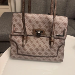 Beige och brun handväska från Guess - Säljer en snygg handväska från Guess i beige med brunt monogrammönster och silverfärgade metalldetaljer. Väskan har två handtag och ett dekorativt lås framtill. Materialet är en blandning av polyester och bomull.