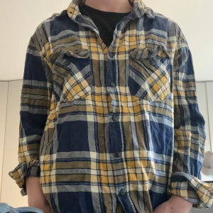 Flanell skjorta  - Säljer en blå och gul rutig skjorta med knappar framtill och två bröstfickor. Skjortan har lång ärm och klassisk krage, perfekt för en avslappnad stil.